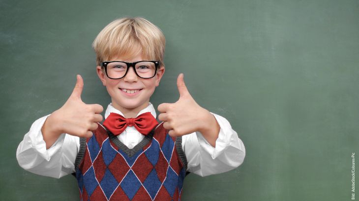 Junge mit Brille zeigt vor einer Schultafel beide Daumen nach oben – Symbolbild zum Schulstart und Versicherungsschutz für Kinder.